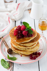 Stack of homemade little pancakes with honey, fresh raspberries and red currants on an old  light wooden background. Selective focus.