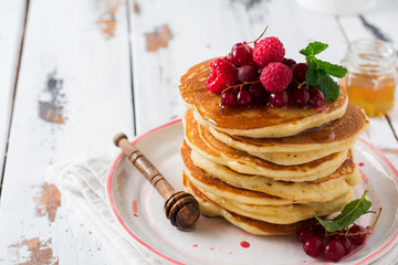 Stack of homemade little pancakes with honey, fresh raspberries and red currants on an old  light wooden background. Selective focus.