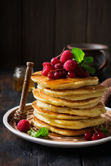 Stack of homemade little pancakes with honey, fresh raspberries and red currants on an old wooden background. Selective focus.