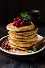 Stack of homemade little pancakes with honey, fresh raspberries and red currants on an old wooden background. Selective focus.