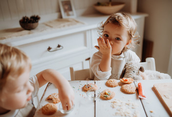 Two small toddler children sitting at the table, decorating and eating cakes at home.