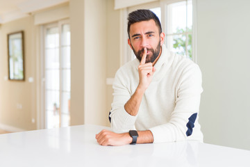 Naklejka premium Handsome hispanic man wearing casual white sweater at home asking to be quiet with finger on lips. Silence and secret concept.