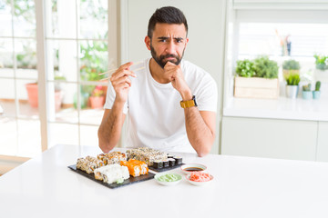 Handsome hispanic man eating asian sushi using chopsticks serious face thinking about question, very confused idea