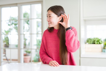 Obraz premium Beautiful Asian woman wearing pink sweater on white table smiling with hand over ear listening an hearing to rumor or gossip. Deafness concept.