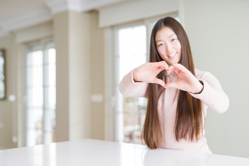 Beautiful Asian woman wearing casual sweater on white table smiling in love showing heart symbol and shape with hands. Romantic concept.