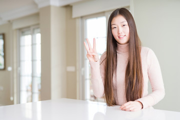 Beautiful Asian woman wearing casual sweater on white table showing and pointing up with fingers number three while smiling confident and happy.