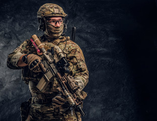 Fully equipped soldier in camouflage uniform holding an assault rifle. Studio photo against a dark textured wall