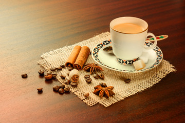 Coffee in a porcelain cup with a saucer, cinnamon, nutmeg and star anise on a jute napkin. Vintage coffee set on wooden table with scattered coffee beans.