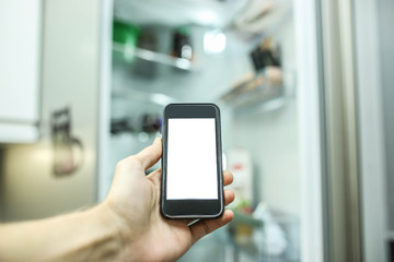 Smartphone with a white screen on the blurred background of an open refrigerator with products in the kitchen. Smart home technology system.