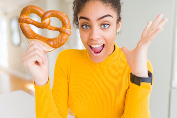 Young african american woman eating German salty pastry pretzel very happy and excited, winner expression celebrating victory screaming with big smile and raised hands