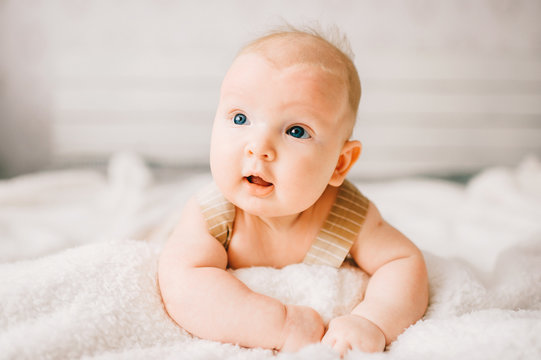 Adorable lovely newborn male baby boy with smiling emotional  happy face lifestyle indoor portrait. Funny infant child lying on stomach on bed with white wall on background.  Carefree childhood.