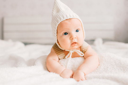 Adorable lovely newborn male baby boy with smiling emotional  happy face lifestyle indoor portrait. Funny infant child in hat lying on stomach on blanket on bed with white wall on background.