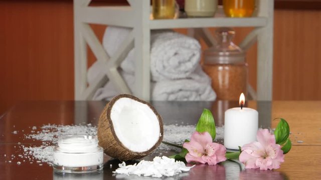 Burning candle, half of coconut , flakes of coconut, cream in a glass cup, pink flowers  are lying on a black  table against the background of the accessories  and cosmetics for spa treatments and aro