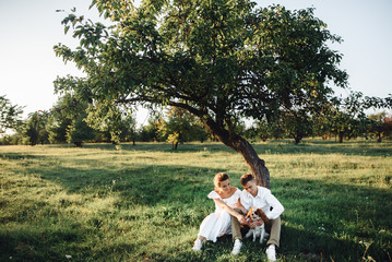 Happy young spouses spend a fun weekend . They sit near a tree on the grass in a park with their little funny dog