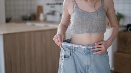 Beautiful female body with measuring tape. Excellent weight loss results. Girl measuring waist with white ribbon. Young woman in living room measures the waist after successful diet.