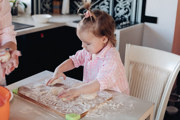 Portrait of lovable child with tail in pink checkered shirt helping mother cooking dumplings or piers in the kitchen. Sitting on chair and kneading dough on board with flour.