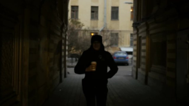 Front view  of young man walking down the city street and drinking coffee. Serious tired man in black walks on the street. 