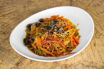 Noodles with vegetables. Mushrooms, zucchini, peppers, carrots in a sweet-sour sauce. Sprinkled with white and black sesame seeds. In a white plate on a wooden background.