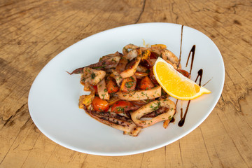 Squid with vegetables and sauce. Sprinkled with fresh dill. Garnish with a slice of lemon. In a white plate on a wooden background.