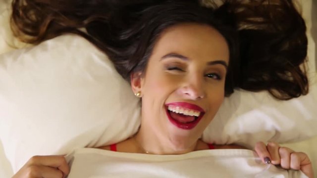 Young attractive woman with cunning look hiding face under white blanket at bedroom. Shy beautiful lady peeking from white duvet. Happy playful wife waiting husband in bed. Top view. Close up video