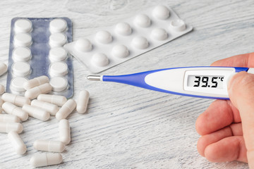 White-blue thermometer with a high temperature of 39.5 degrees Celsius in hand with pills and medical capsules