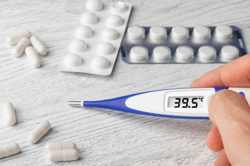 White-blue thermometer with a high temperature of 39.5 degrees Celsius in hand with pills and medical capsules