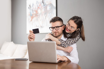 Young happy married couple sitting at table in apartment and holding credit card