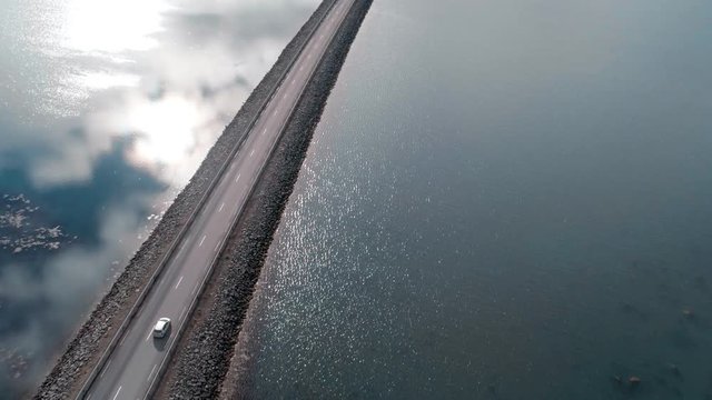 Aerial drone shot of endless infinity road going into horizon on clear summer day, bridge connects two banks of lake or bay, crystal clear water simmers in soft breeze or wind. Small car drives by