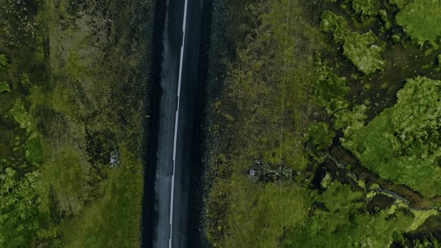 Straight down shot of drone following turns and curves of winding mountain road through contrasted and colorful bright green fields of moss in iceland landscape. Wet tarmac asphalt surface