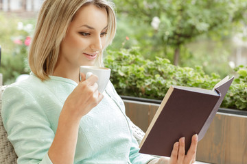What a pleasant morning. Beautiful young woman smiling and reading a book holding a cup of coffee