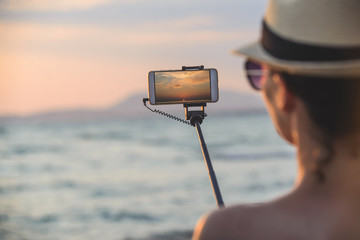 Beautiful young woman makes landscape photo for Instagram at sunset by the sea with selfie stick