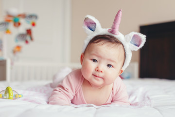 Portrait of cute adorable Asian mixed race smiling baby girl four months old lying on tummy on bed in bedroom wearing unicorn headband horn and ears looking in camera. © anoushkatoronto