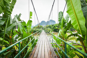 Fototapety  Wooden suspension bridge crossing the river to tropical forest with banana leaf plant