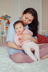 Portrait of beautiful mixed race Asian mother holding touching embracing her cute adorable newborn infant baby. Early development and health care lifestyle concept. Family in bedroom