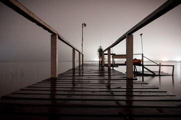 Silhouette of a man standing on a wooden dock in the dark staring at the emptiness of a lake with glowing lights on the opposite side