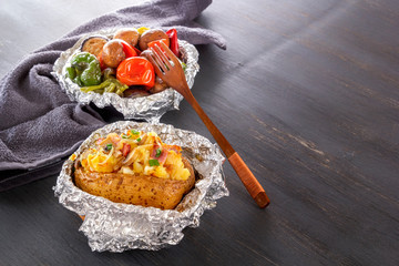 Baked potatoes with bacon, onions and baked vegetables in foil - tomatoes, eggplants, peppers on a gray wooden table.