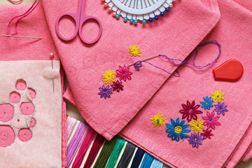 Needlework. Close-up view to embroidery process of colorful flowers on pink kitchen napkins. Making...