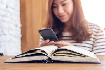 Closeup image of a beautiful asian woman holding , using and looking at smart phone with a book on wooden table