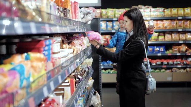 Beautiful woman in glasses holding her child in her arms while choosing baby food on the shelves in the supermarket. houghtful mother looks at the goods, carefully selects goods for her child.