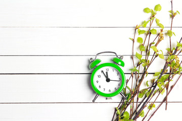 Green alarm clock and young birch branches with first leaves and buds on a white wooden background.