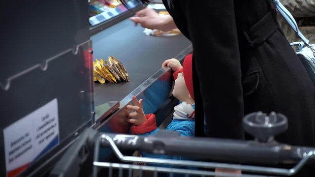 A child of three years old, warmly dressed, stands next to the supermarket cashier, looks at the goods and begins to cry