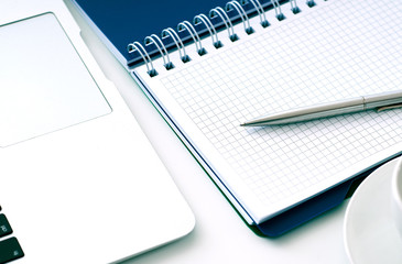 Closeup of white desktop with laptop, coffee cup, notepads, pen, calculator and other items.