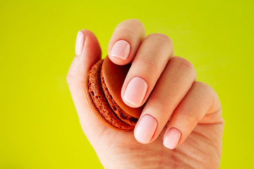 Hand with light rose manicure with brown chocolate macaron. Isolated at bright yellow background.