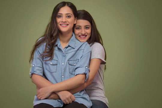 Studio Shot Of Two Smiling Young Teenager Girls Sitting In A Room Holding Each Other	