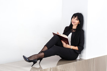 Young Beautiful happy brunette woman holding a notebook dressed in black business suit sitting on a floor in office, smiling, looking at camera.