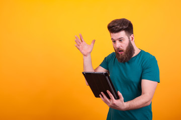 Handsome bearded man in green t-shirt looking shocked at tablet over yellow background