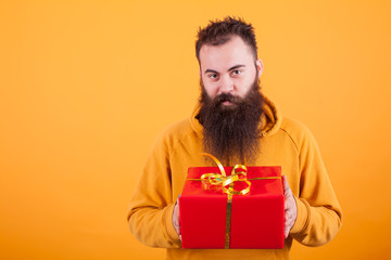 Cheerful young man with long beard holding a red gift box over yellow background