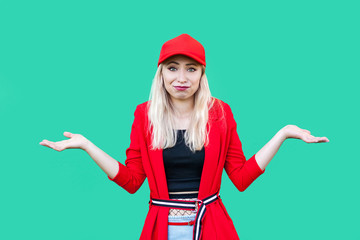 I don't know. Portrait of confused beautiful blond young hipster style woman in red blouse and cap, standing with raised arms and looking at camera. indoor studio shot, isolated on green background.