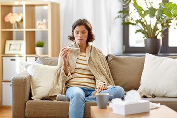 people, health and fever concept - sad sick woman in blanket measuring temperature by thermometer at home