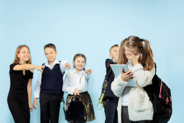 Little girl standing alone and suffering an act of bullying while children mocking. Sad young schoolgirl sitting on studio against blue background.